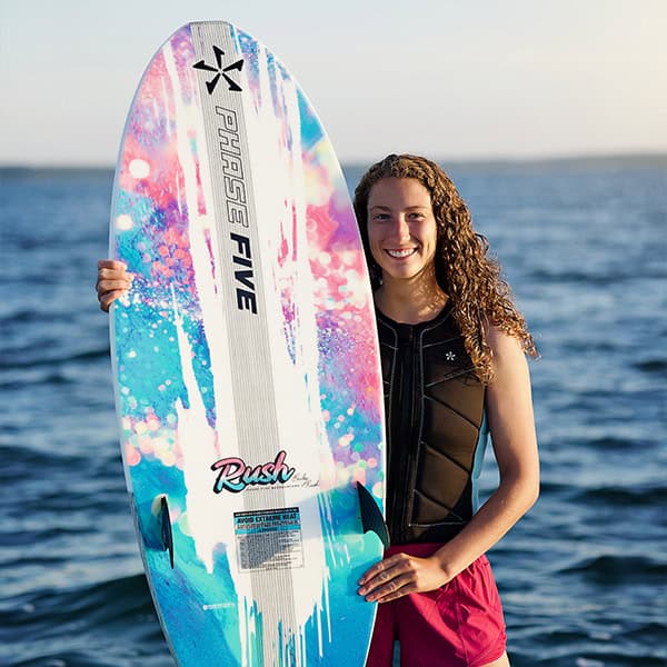 A woman smiling and holding a vibrant, colorful surfboard near the ocean, ready for surfing. The surfboard features a mix of pink, blue, and white colors with branding and design elements.