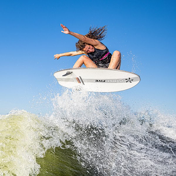 A woman with curly hair jumping on a surfboard above a wave under a clear blue sky.